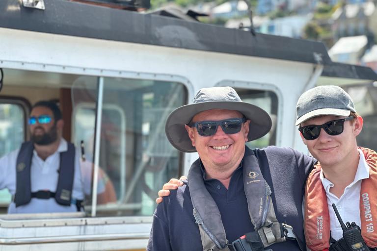 A photograph of staff working on the Dartmouth lower ferry. In the foreground, two attendants wearing hats, sunglasses and safety gear are smiling at the camera, while in the background, the skipper can be seen in the cab  steering the ferry