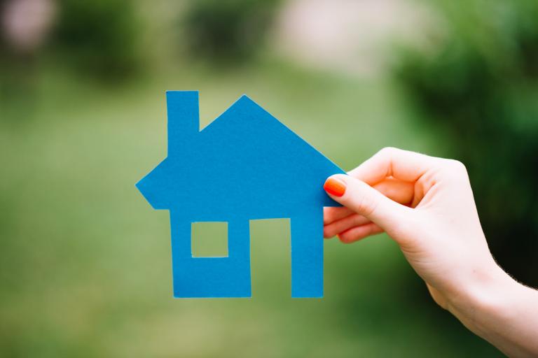 A hand holding a blue house made of paper with a blurred green woodland in the background.