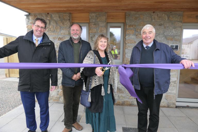 Four people stand in a line, about to cut a large purple ribbon to unveil new housing.