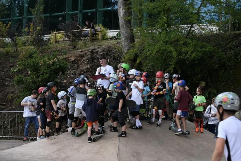 Large group of children gather around adult giving instructions on skate ramp