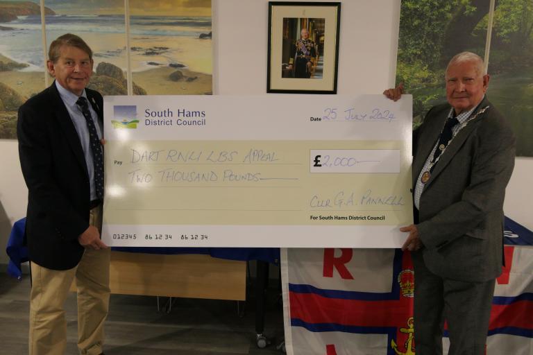 Councillor Guy Pannell, vice chair of South Hams District Council, pictures presenting a cheque to Commodore Jake Moores, chairman of the Dart RNLI Appeal. The cheque is an oversized novelty cheque with the council logo on it. Jake and Councillor Pannell are smiling while holding the cheque.