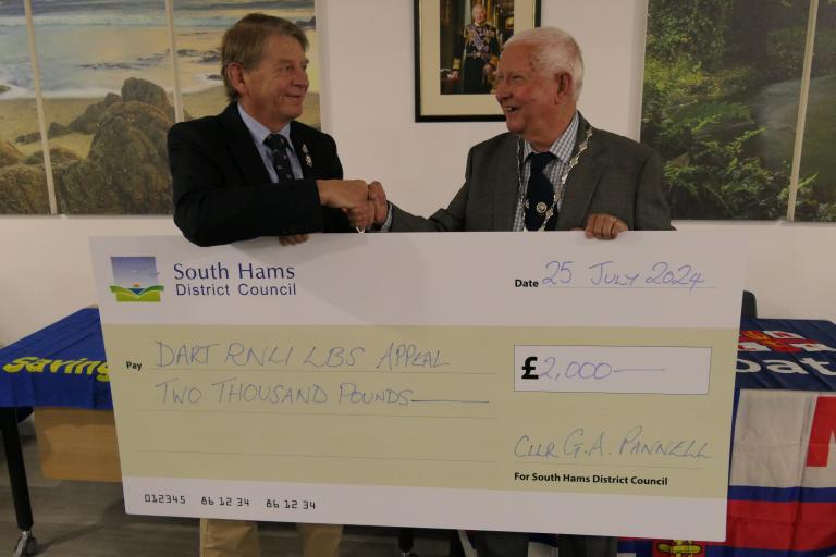 Councillor Guy Pannell, vice chair of South Hams District Council, pictures presenting a cheque to Commodore Jake Moores, chairman of the Dart RNLI Appeal. The cheque is an oversized novelty cheque with the council logo on it. Jake and Councillor Pannell are smiling and shaking hands while holding the cheque.