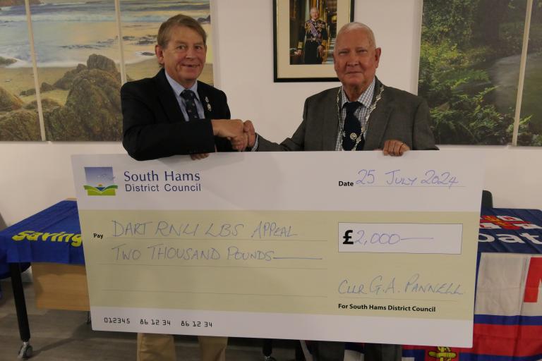 Councillor Guy Pannell, vice chair of South Hams District Council, pictures presenting a cheque to Commodore Jake Moores, chairman of the Dart RNLI Appeal. The cheque is an oversized novelty cheque with the council logo on it. Jake and Councillor Pannell are smiling and shaking hands while holding the cheque.