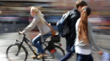 A blonde woman is riding a bicycle in a town centre, heading from right to left. A young couple, both with brown hair, walk in the opposite direction. They are deep in discussion.