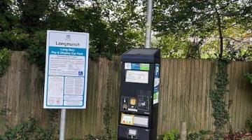 Car park pay and display machine with yellow pay here above and a white sign to the left with information about parking charges on. Autumn leaves fallen on the floor and a wooden fence behind with trees at the back.