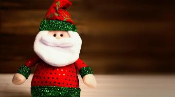 A plush cuddly toy Father Christmas sat on a plain wooden table, with a plain burgundy background.