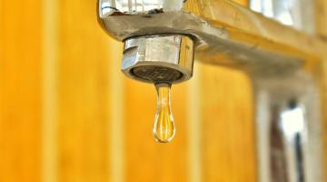 A drop of water pouring from a tap. The tap is silver, and the wall behind it is bright yellow.