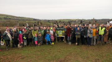 A group of people stand around a sign which says Hope Wood in a field, with rolling hills in the background.