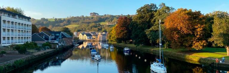 A river with small boats on it. A white building to the riverbank on the left with trees to the bank on the right. Trees are in the distance and the sky is clear blue. 